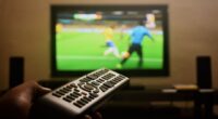 Controle remoto apontado para televisão exibindo partida de futebol, representando a preferência dos belo-horizontinos por acompanhar os jogos da Copa do Mundo em casa