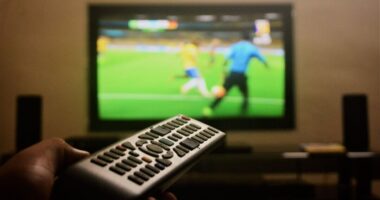 Controle remoto apontado para televisão exibindo partida de futebol, representando a preferência dos belo-horizontinos por acompanhar os jogos da Copa do Mundo em casa