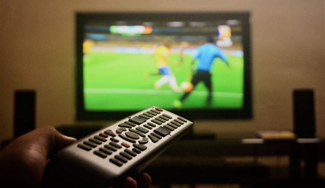 Controle remoto apontado para televisão exibindo partida de futebol, representando a preferência dos belo-horizontinos por acompanhar os jogos da Copa do Mundo em casa Controle remoto apontado para televisão exibindo partida de futebol, representando a preferência dos belo-horizontinos por acompanhar os jogos da Copa do Mundo em casa