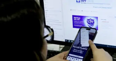 Pessoa utiliza aplicativo da Receita Federal em smartphone enquanto acessa informações sobre a declaração do Imposto de Renda 2026 em um computador para consultar a restituição do imposto