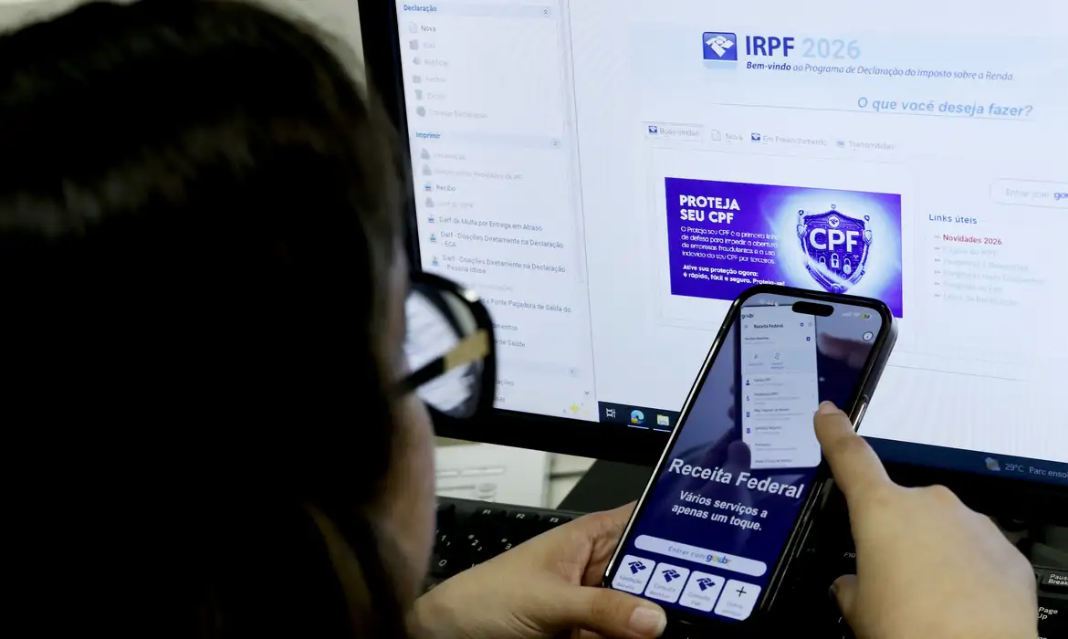 Receita Federal libera consulta ao segundo lote da restituição do Imposto de Renda 2026 Pessoa utiliza aplicativo da Receita Federal em smartphone enquanto acessa informações sobre a declaração do Imposto de Renda 2026 em um computador para consultar a restituição do imposto