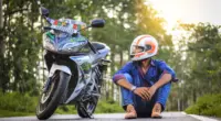 Motociclista com capacete sentado ao lado de uma motocicleta estacionada em uma estrada arborizada, imagem que simboliza o Dia do Motociclista e a importância da segurança sobre duas rodas