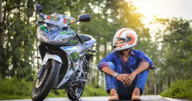 Motociclista com capacete sentado ao lado de uma motocicleta estacionada em uma estrada arborizada, imagem que simboliza o Dia do Motociclista e a importância da segurança sobre duas rodas