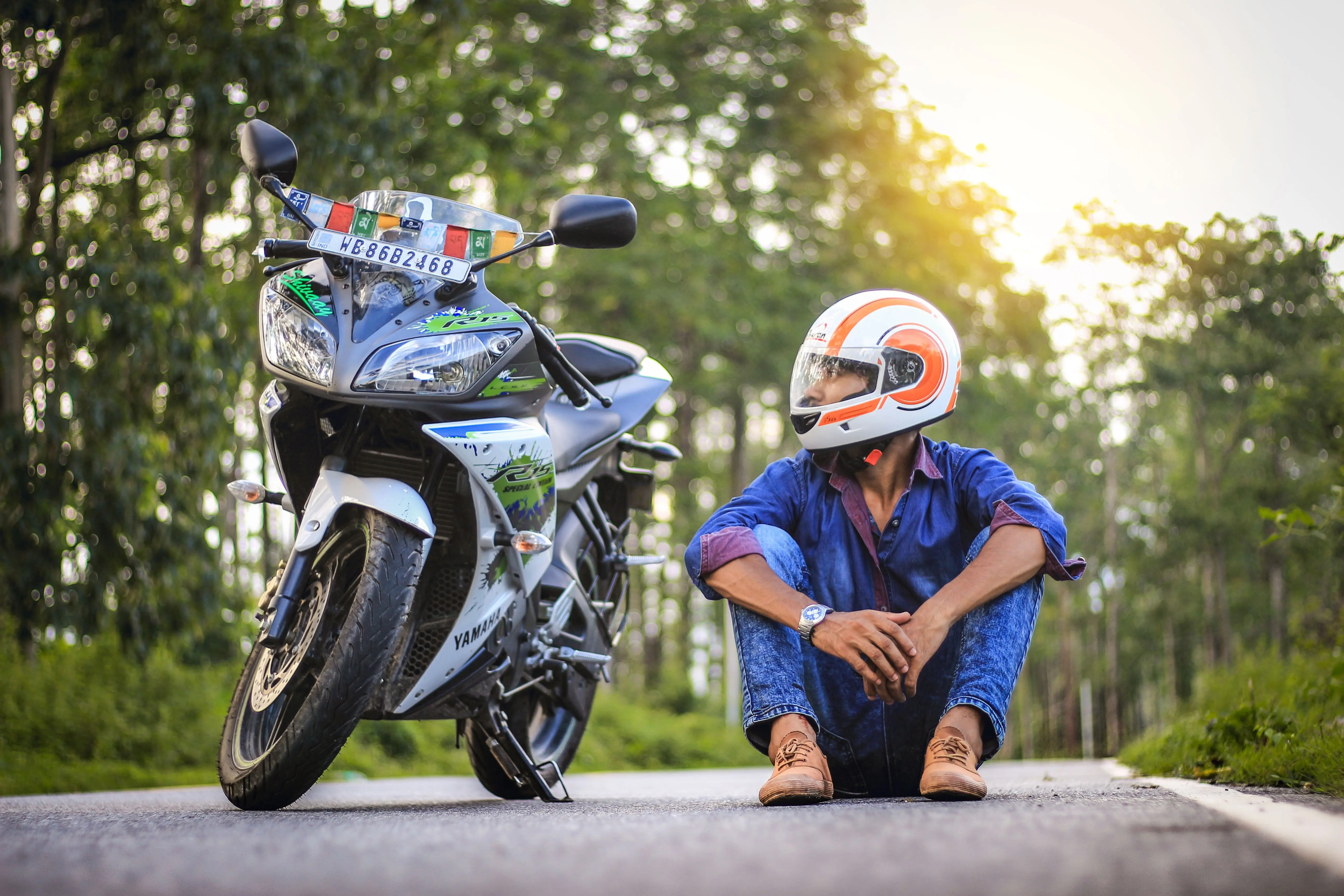 Dia do Motociclista Motociclista com capacete sentado ao lado de uma motocicleta estacionada em uma estrada arborizada, imagem que simboliza o Dia do Motociclista e a importância da segurança sobre duas rodas