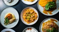 Pratos variados dos sabores brasileiro, com massas, carnes, frutos do mar e acompanhamentos servidos em louças individuais.
