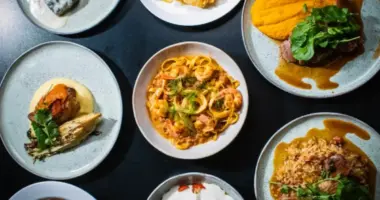 Pratos variados dos sabores brasileiro, com massas, carnes, frutos do mar e acompanhamentos servidos em louças individuais.