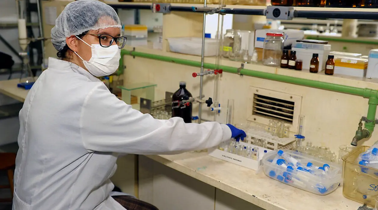 Pesquisadora trabalha em laboratório da UFMG Pesquisadora com jaleco, máscara, touca e luvas manuseia materiais em uma bancada de laboratório da Universidade Federal de Minas Gerais que se destacou em Ranking Internacional