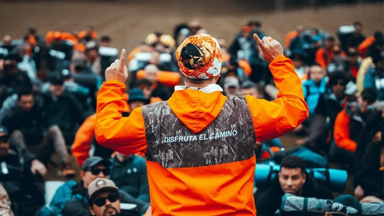 Encontro do movimento Legendários reúne participantes Homem com roupa laranja ergue os braços diante de um grupo de pessoas reunidas em um ambiente fechado durante a jornada dos Legendários